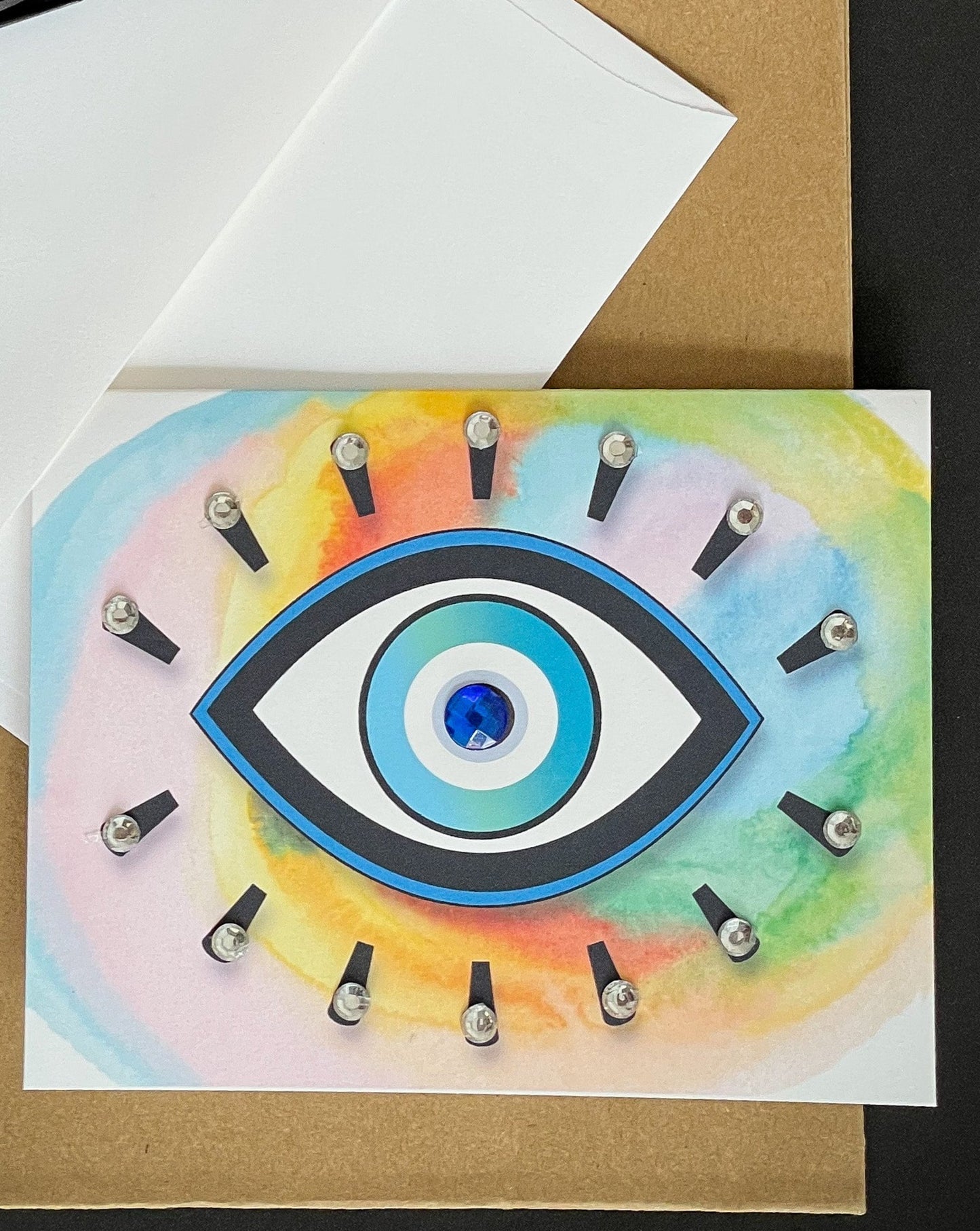 this is a note card on a craft paper gift bag with a white envelope and a black marker , the card has an evil eye symbol on a colourful background