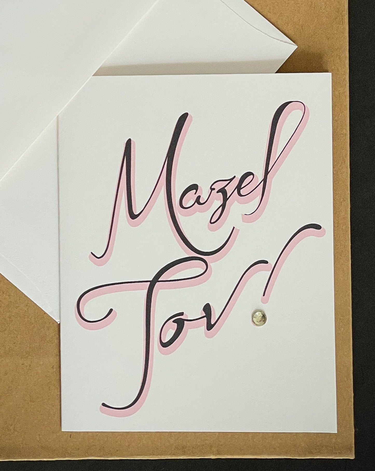 this is a note card on a craft paper gift bag with a white envelope and a black marker , the card says Mazel Tov in black with a pink shadow