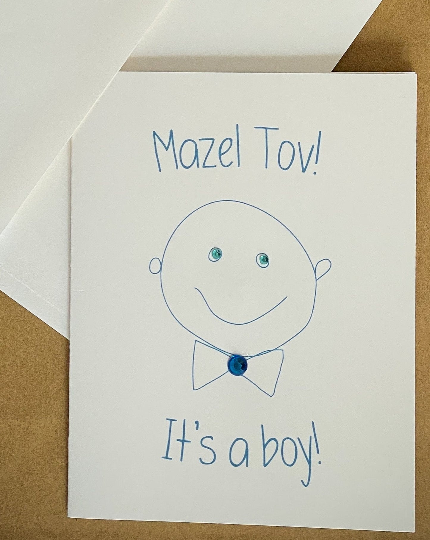 this is a note card on a craft paper gift bag with a white envelope and a black marker , the card says Mazel Tov! It’s A Boy! With a cute drawing of a baby boy