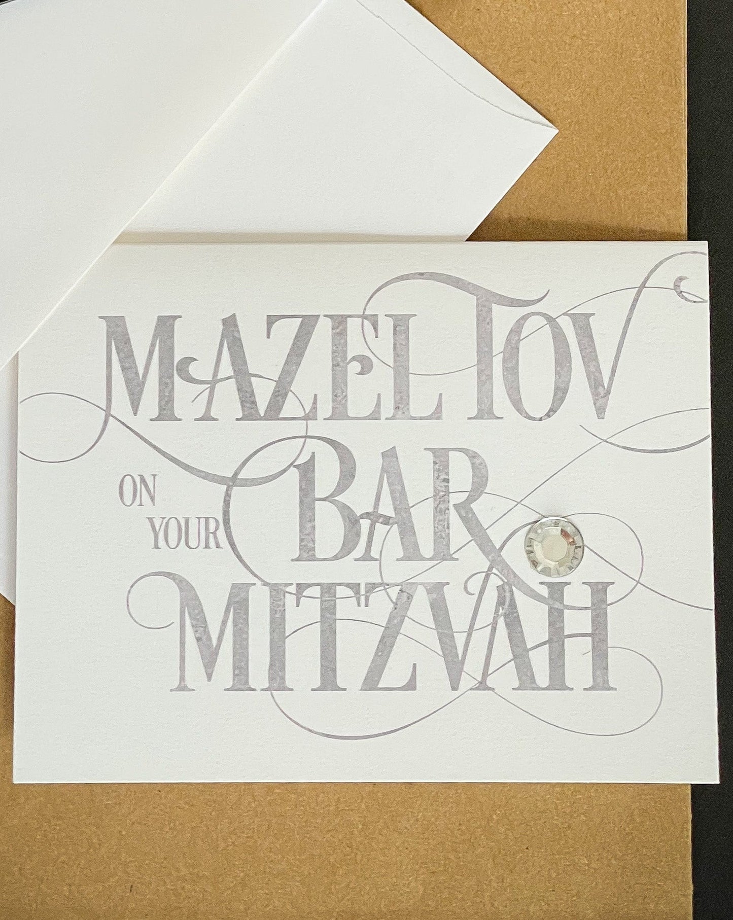 this is a note card on a craft paper gift bag with a white envelope and a black marker , the card says Mazel Tov on your bar mitzvah in a silver script font