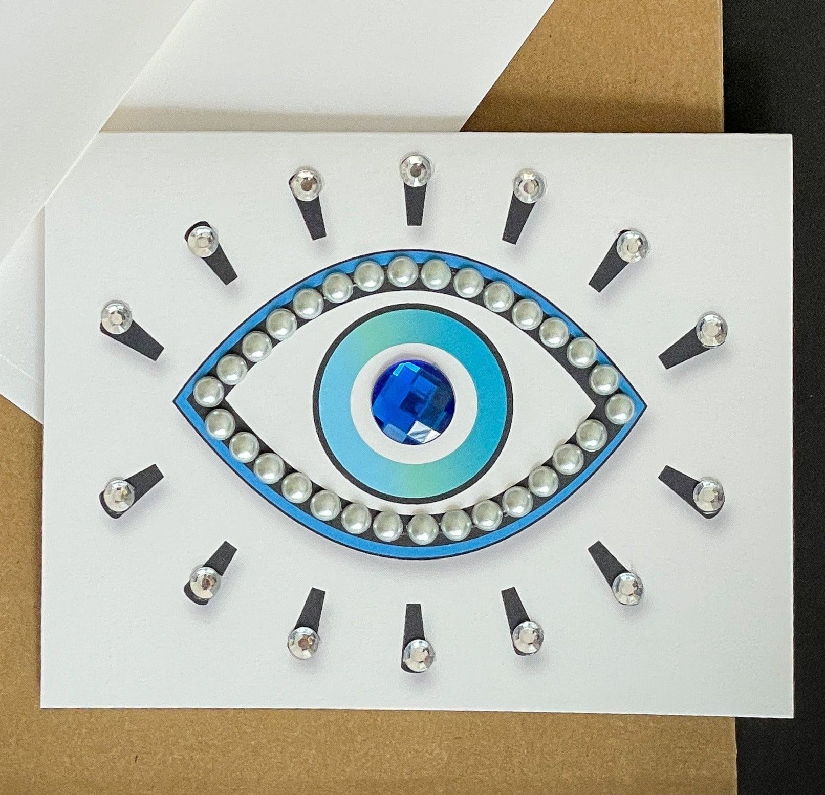 this is a note card on a craft paper gift bag with a white envelope and a black marker , the card has an evil eye symbol decorated with pearls and clear stones