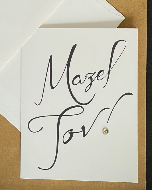 this is a note card on a craft paper gift bag with a white envelope and a black marker , the card says Mazel Tov in black 