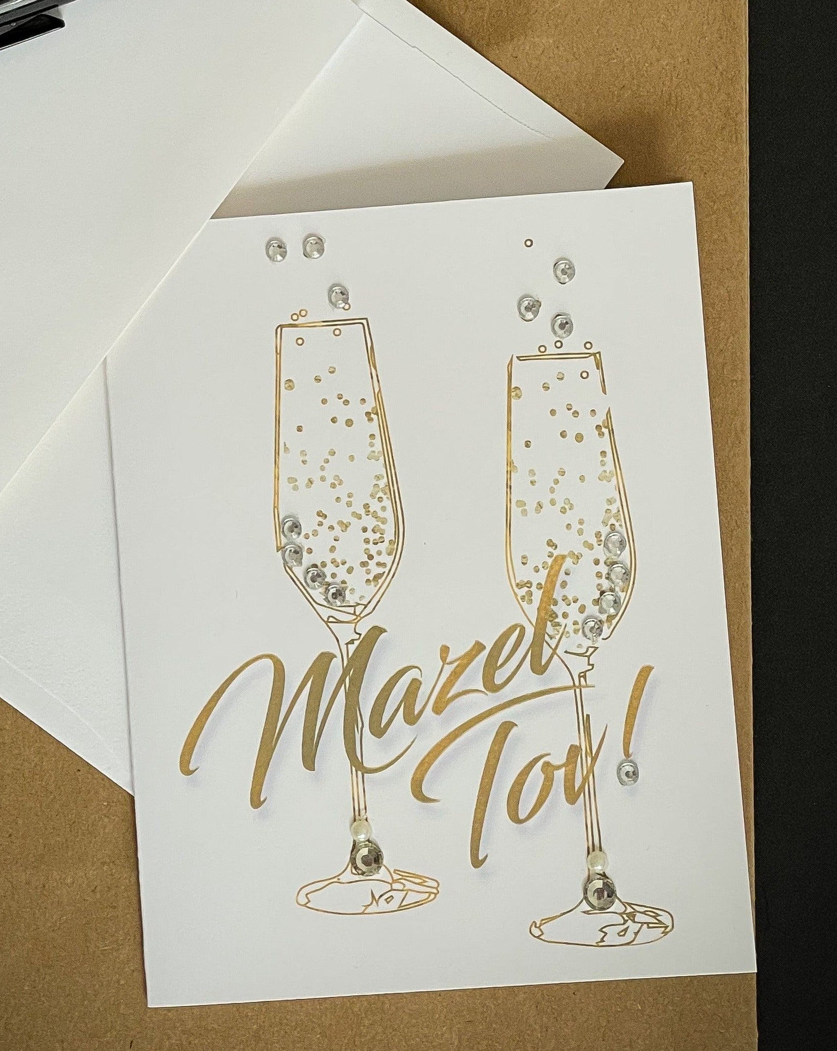 this is a note card on a craft paper gift bag with a white envelope and a black marker , the card says Mazel Tov in gold with champagne flutes 