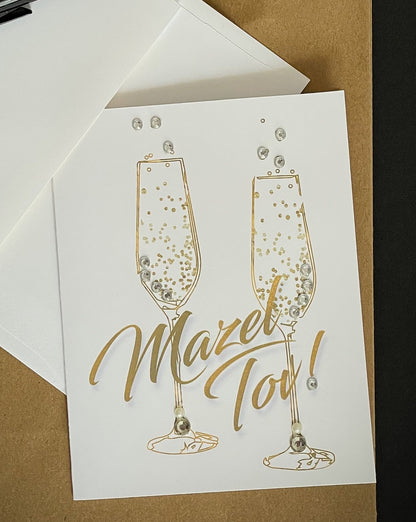 this is a note card on a craft paper gift bag with a white envelope and a black marker , the card says Mazel Tov in gold with champagne flutes 