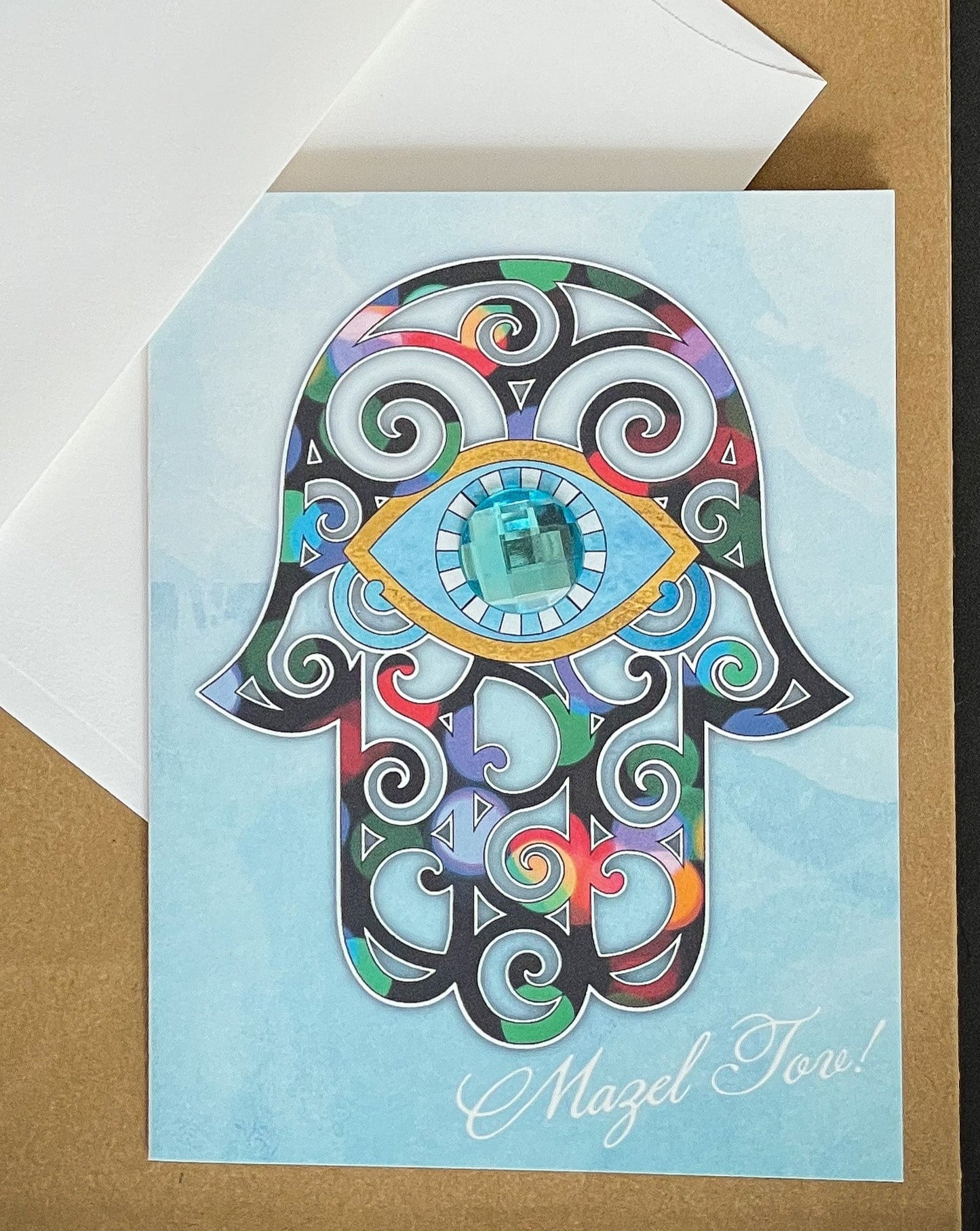 this is a note card on a craft paper gift bag with a white envelope and a black marker , the card has a hamsa on it that looks like stained glass with Mazel Tov written in the bottom right corner