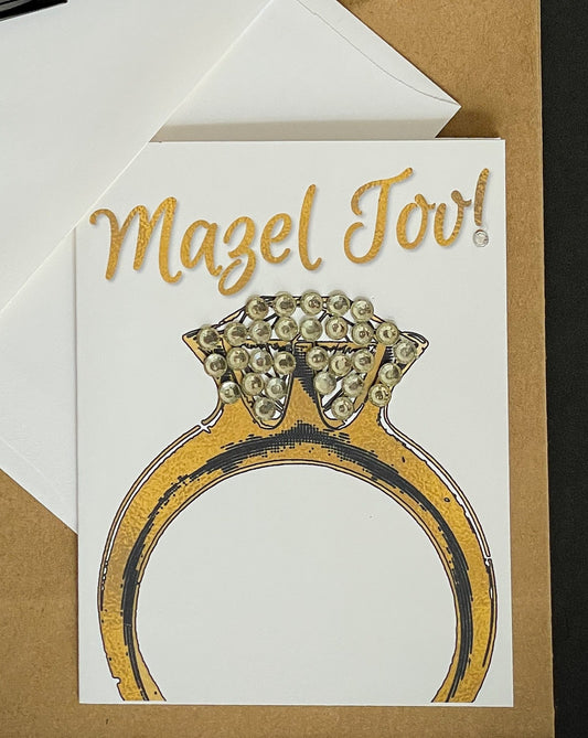 this is a note card on a craft paper gift bag with a white envelope and a black marker , the card says Mazel Tov and has a beautiful engagement ring in gold with jewelled diamond area
