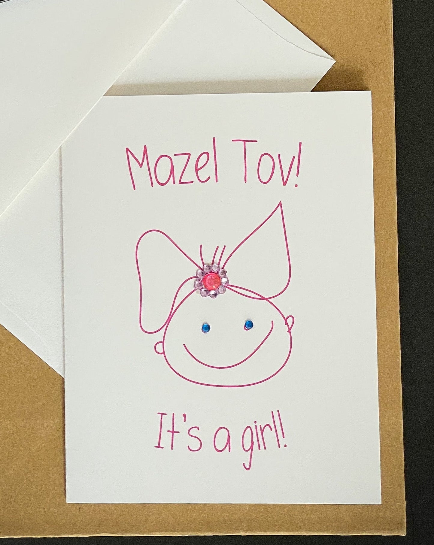 this is a note card on a craft paper gift bag with a white envelope and a black marker , the card says Mazel Tov! It’s A Girl! With a cute drawing of a baby girl