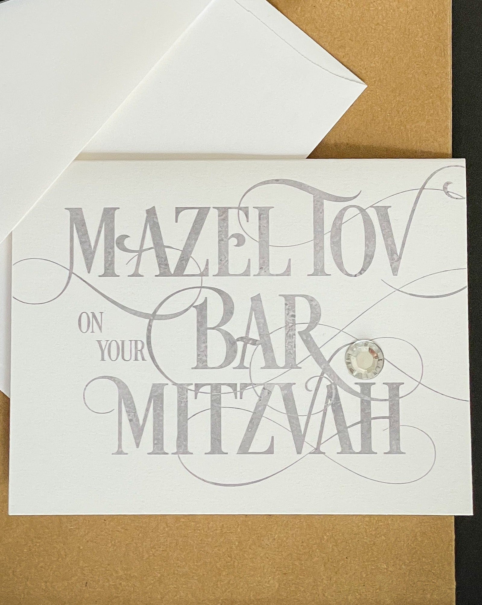this is a note card on a craft paper gift bag with a white envelope and a black marker , the card says Mazel Tov on your bar mitzvah in a silver script font