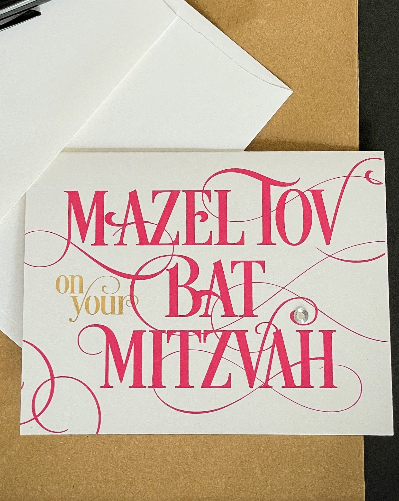 this is a note card on a craft paper gift bag with a white envelope and a black marker , the card says Mazel Tov on your bat mitzvah in a hot pink script font