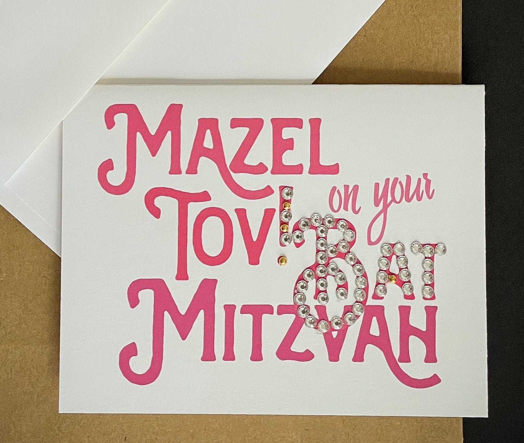 this is a note card on a craft paper gift bag with a white envelope and a black marker , the card says Mazel Tov on your bat mitzvah in pink