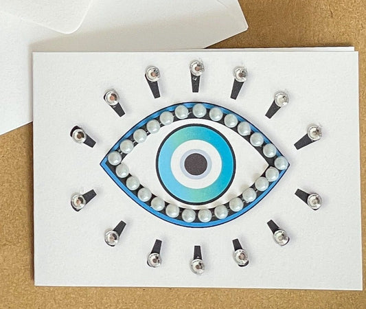 this is a mini note card on a craft paper gift bag with a white envelope and a black marker , the card has an evil eye symbol decorated with pearls and clear jewels