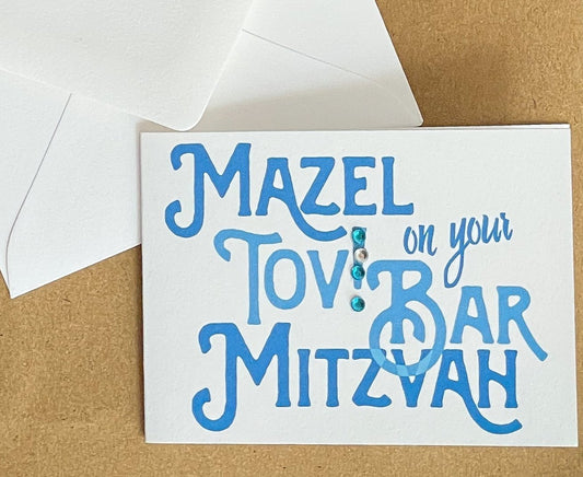 this is a mini note card on a craft paper gift bag with a white envelope and a black marker , the card says Mazel Tov on your bar mitzvah in blue
