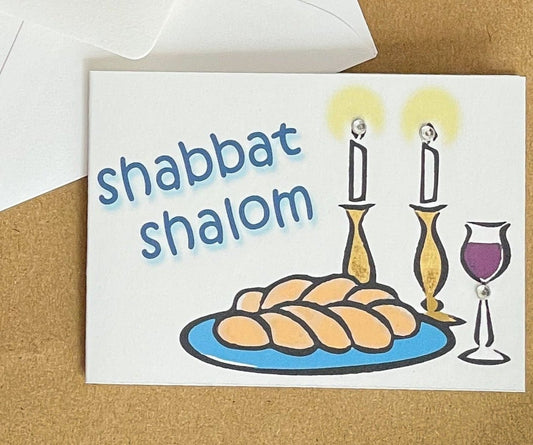 this is a mini note card on a craft paper gift bag with a white envelope and a black marker , the card says Shabbat Shalom with a traditional shabbat set up of challah, wine and candles