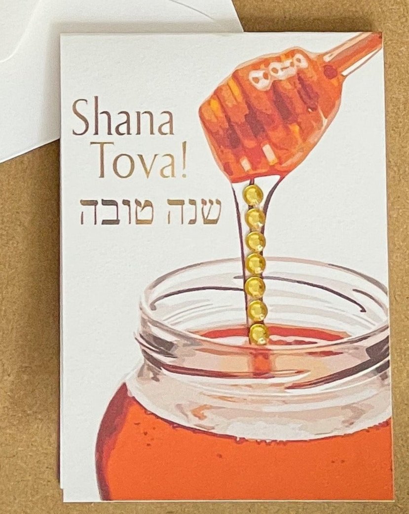 this is a mini note card on a craft paper gift bag with a white envelope and a black marker , the card says Shana Tova in English and Hebrew with a glass jar of honey and honey pouring down from the honey server